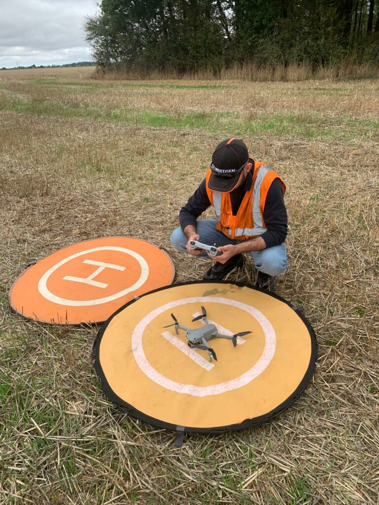 Télépilote MGI accroupi dans un champ près de deux aires d'atterrissage, drone prêt au décollage