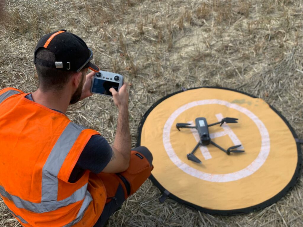 Télépilote MGI en gilet haute visibilité vérifiant sa radiocommande devant un drone posé sur une aire d'atterrissage