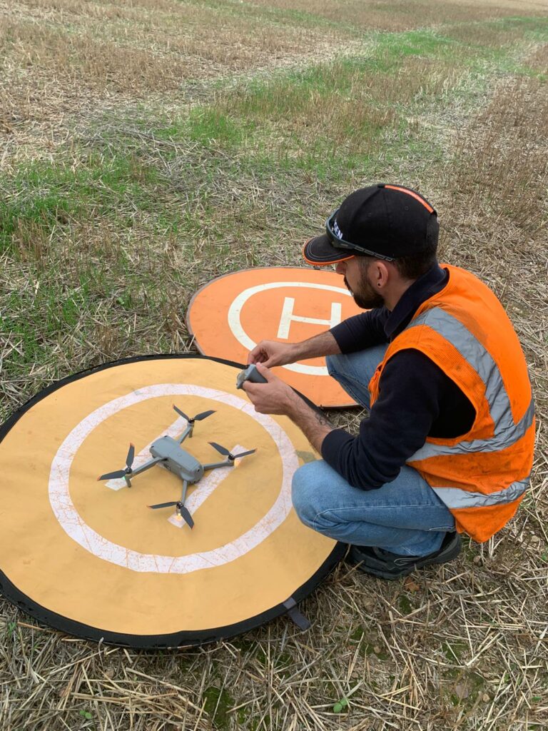 Télépilote MGI en gilet orange contrôlant un drone sur une aire d'atterrissage avant le vol